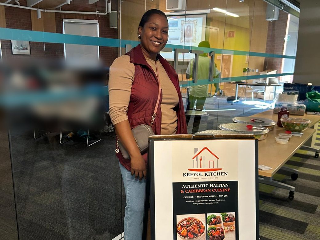 A woman smiles beside a Kreyol Kitchen sign advertising authentic Haitian and Caribbean cuisine, with lunch set out on a table.