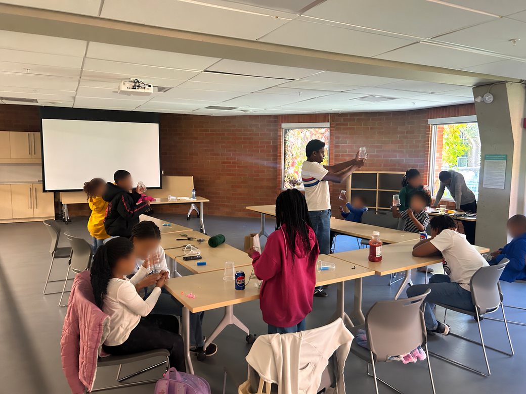 Younger students hold plastic cups to their ears while testing the sound amplifiers they built, as an instructor demonstrates at the front of the room.